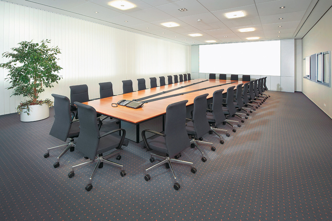 Aufnahme eines kompletten, hellen Konferenzraumes mit langem Tafel-Tisch, schwarzen Drehstühlen und Zimmerpflanze in der linken Ecke.